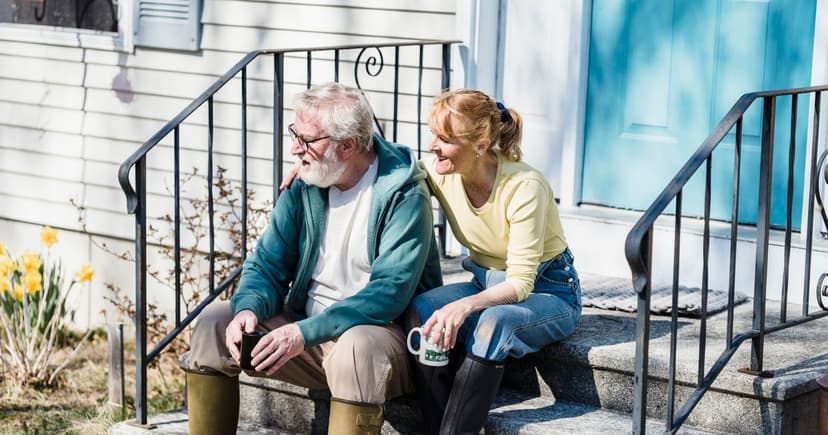 A man in a green zip up sweatshirt and a woman in a yellow top sit close together on the granite steps of a white clapboard house, both in rubber boots and holding mugs of coffee, with daffodils out beside the railing.