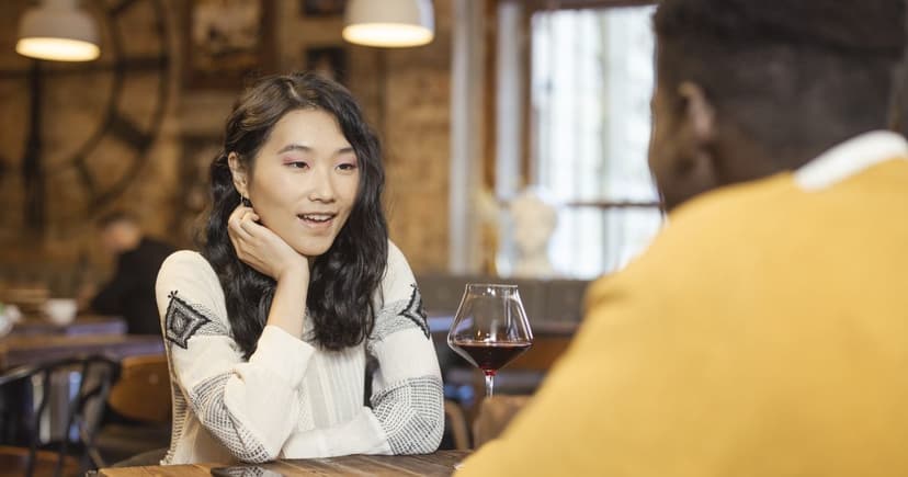 A woman in a cream patterned sweater leans on her hand across a wooden table from a man in a mustard sweater, a glass of red wine between them in a brick walled room.