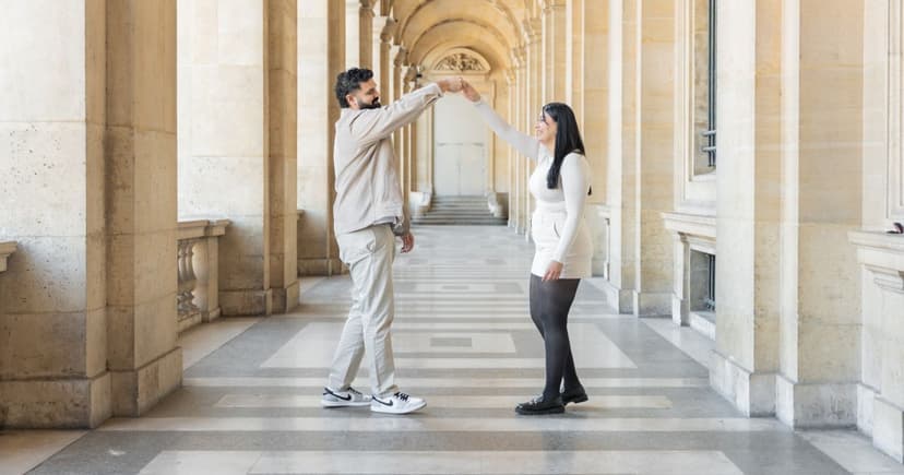 A man and a woman hold hands overhead mid twirl in a sunlit limestone colonnade, both dressed in cream and white.