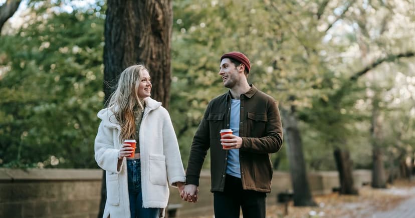 A smiling couple in warm coats walk hand in hand along a leafy park path, each holding a red takeout coffee cup.