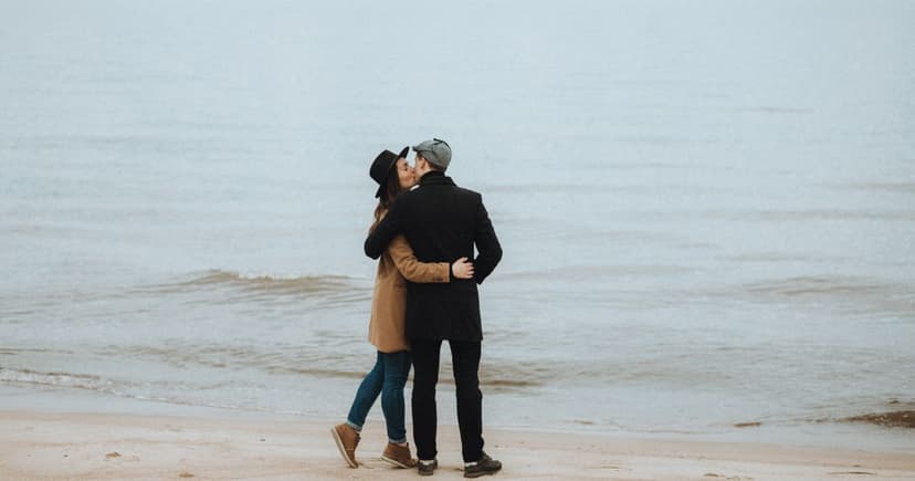 A couple in wool coats and hats stand together on wet sand at the edge of a misty grey sea.