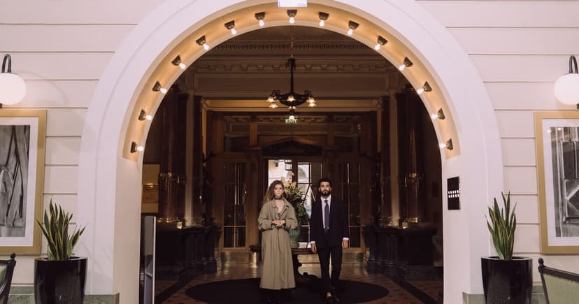 A woman in a long trench coat and a man in a dark suit stand under the lit arch of a grand hotel entrance.
