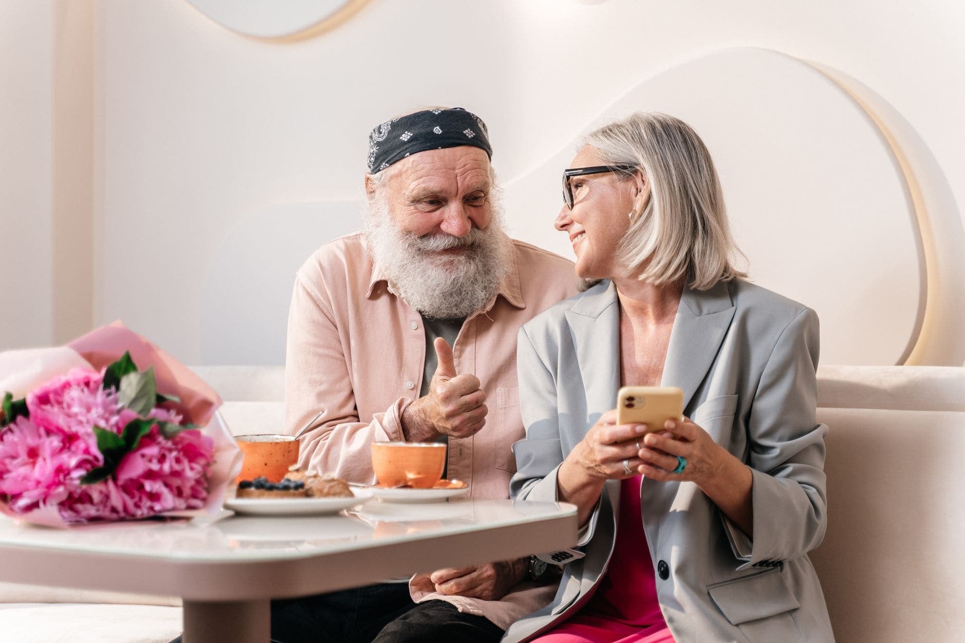 Duas pessoas sentam-se à mesa de um café com chávenas; uma segura um telemóvel e sorri para a outra.