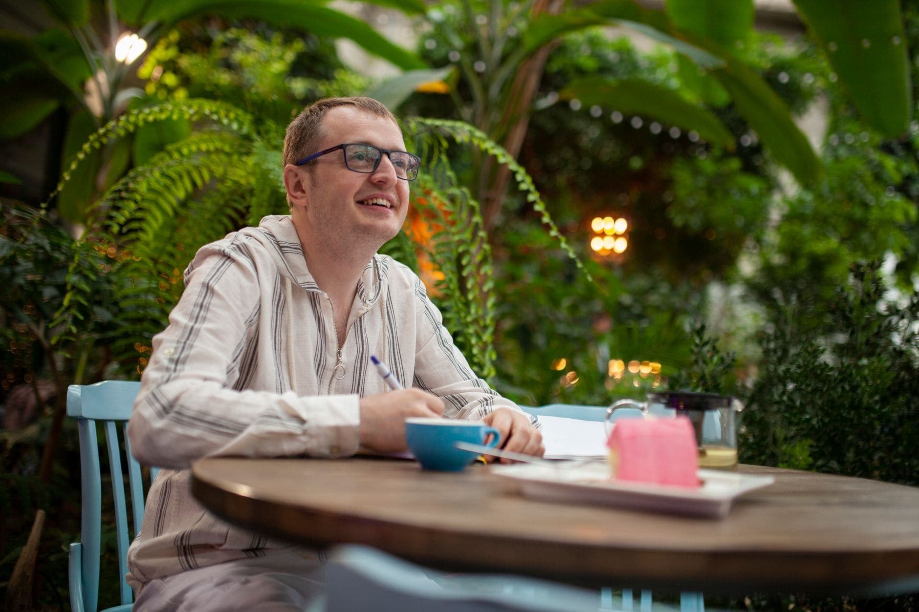 A man with glasses holds a pen above a notebook at a café table among green plants.