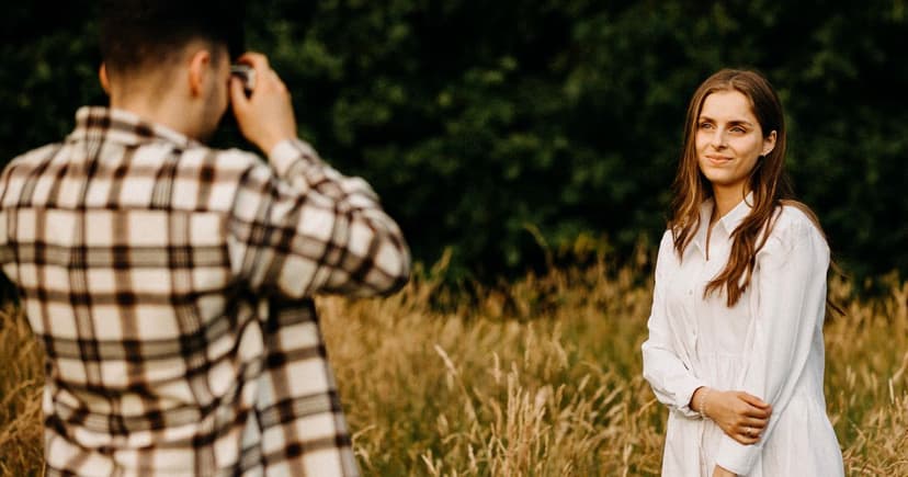 A man in a checked shirt photographs a smiling woman standing in a sunlit grassy field in warm evening light