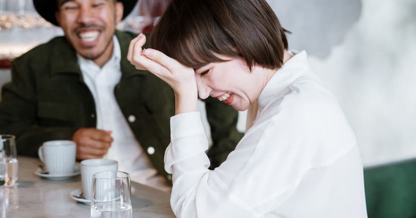 A woman in a white shirt laughs with her hand to her forehead while a man across the cafe table laughs with her over coffee