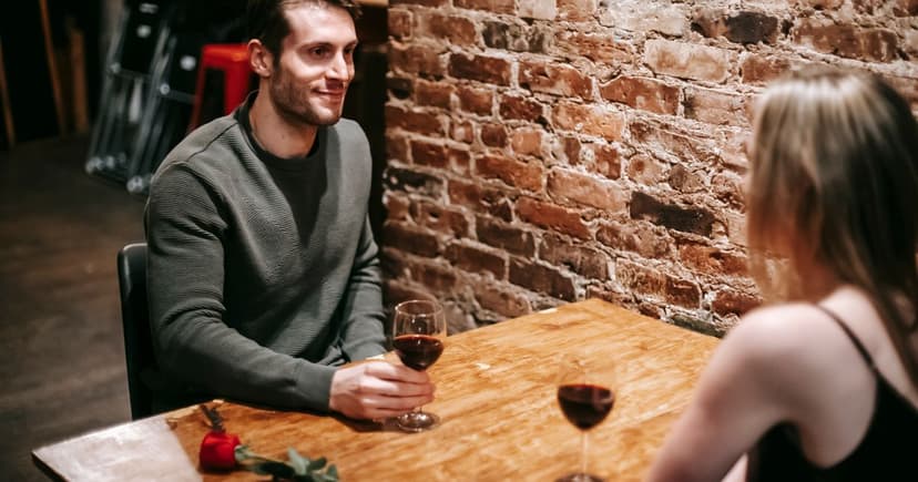 A smiling man holding a glass of red wine across the table from his date in a cosy brick-walled wine bar