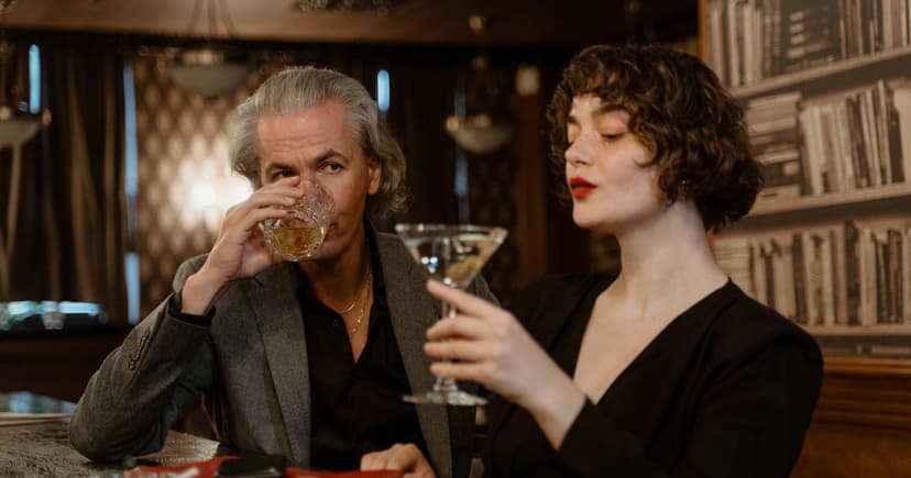 Ein Mann mit grauem Haar und eine jüngere Frau sitzen mit Whisky und Cocktail nebeneinander an einer holzvertäfelten Bar.
