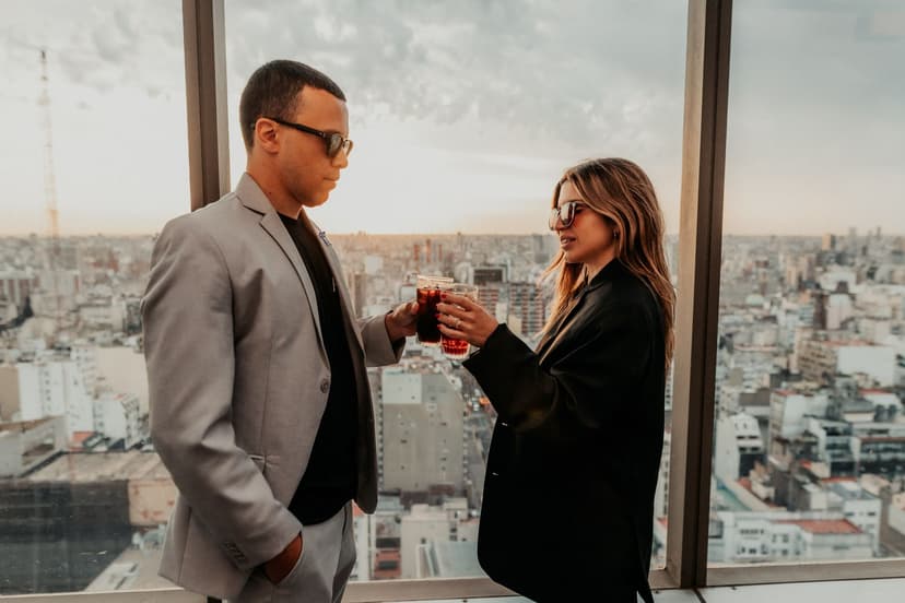 A stylish couple in blazers toast with iced drinks in front of floor-to-ceiling windows overlooking a sprawling city skyline at sunset.