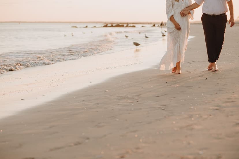 An elegant couple walks hand in hand along the wet sand at golden hour, her long white dress catching the breeze as seagulls gather at the water's edge.