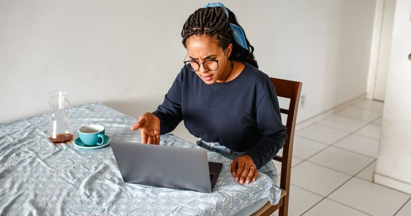 A woman at her kitchen table frowning sceptically at her laptop screen beside a cup of coffee