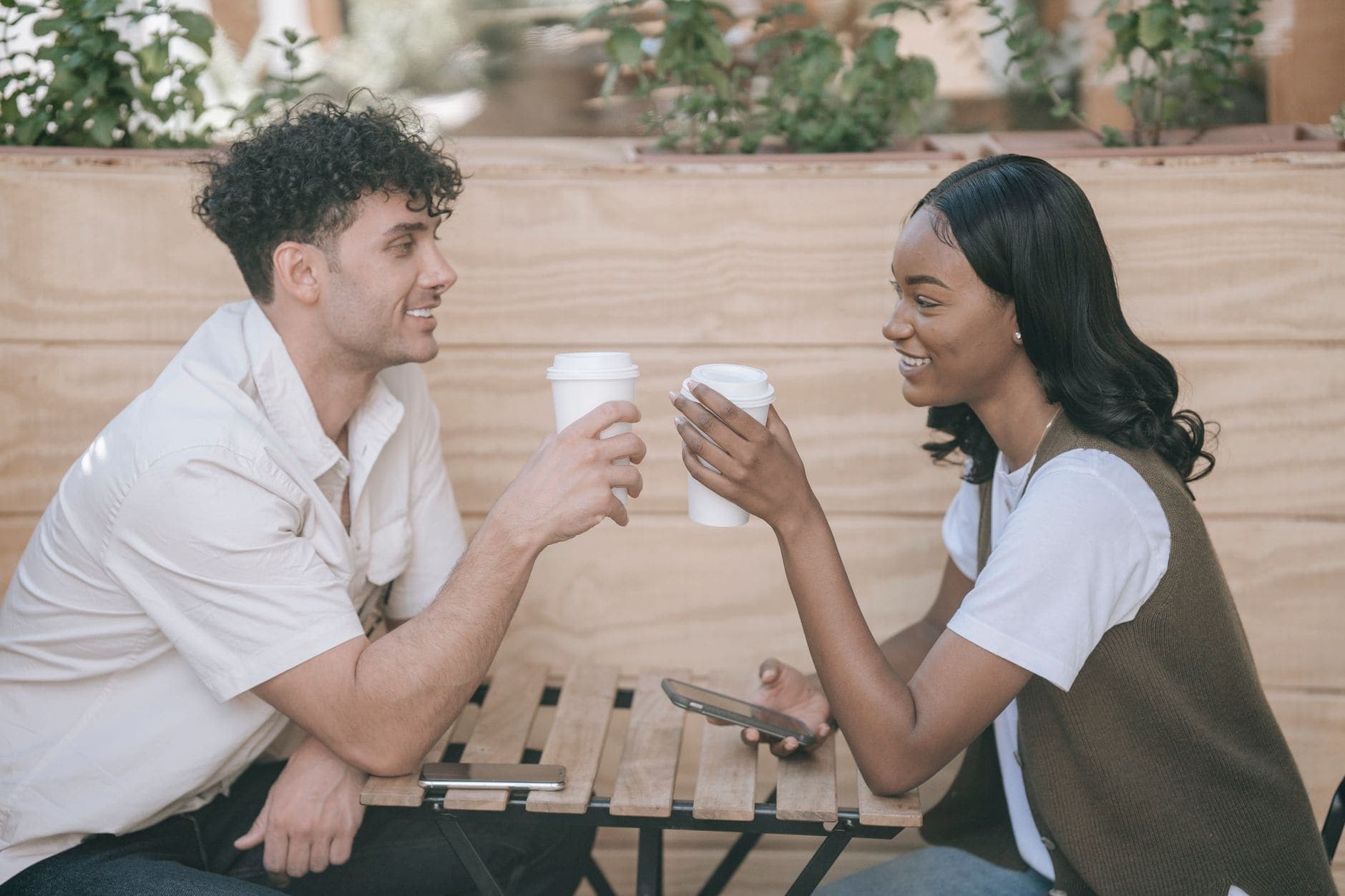 Two adults smile at each other over white takeaway cups at a wooden café table.