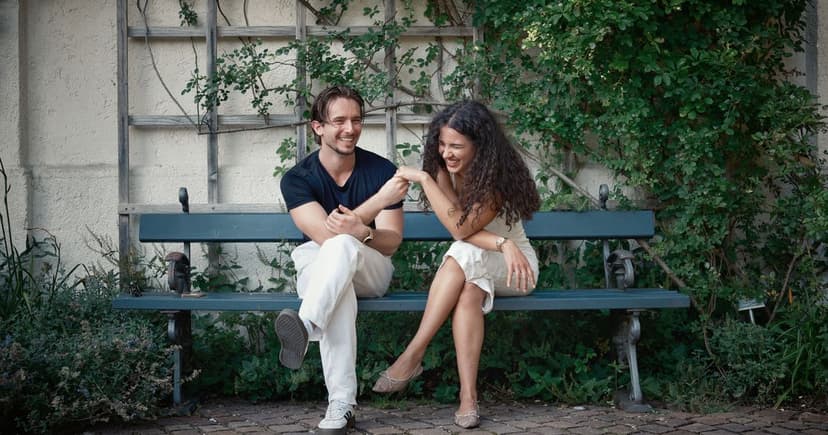A laughing couple in linen sits on a green park bench against a plaster wall covered with climbing greenery.