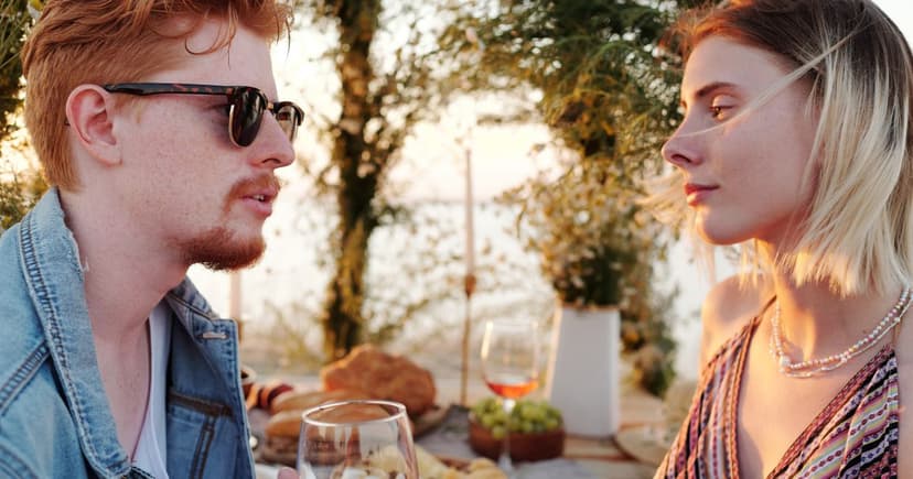 A man in denim and a blonde woman face each other over wine glasses at an outdoor table in golden evening light.
