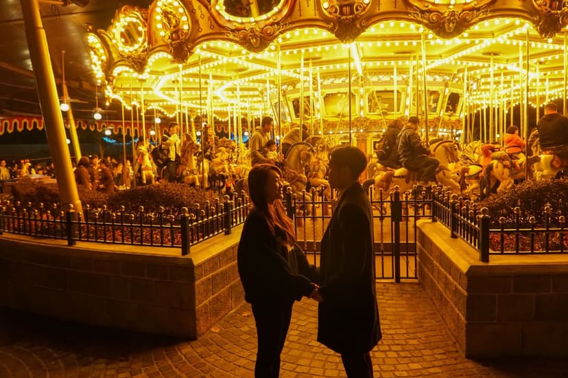 Well-dressed couple holding hands and smiling at each other in front of a glowing carousel at an evening fair, bathed in warm golden light