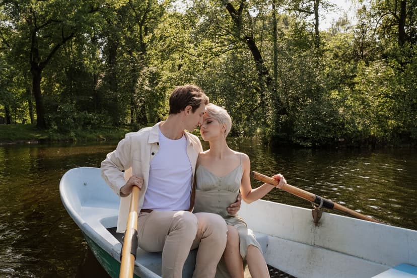 Stylish young couple sharing a tender moment in a rowboat on a calm tree-lined lake, each resting a hand on an oar in the summer sunlight