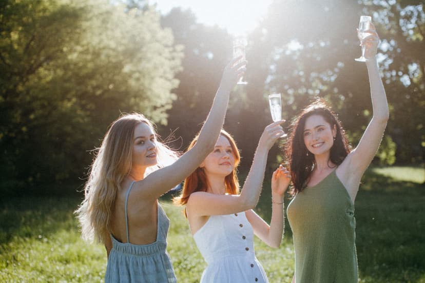 Three women in elegant summer dresses raising champagne flutes in golden sunlight at a garden party, with trees behind them