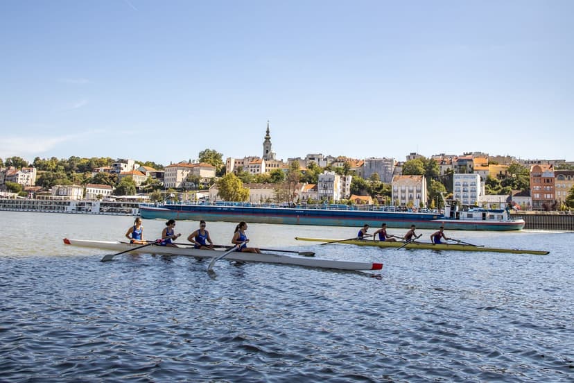 Two four-person rowing crews racing side by side on a sunlit river, with an old town skyline and a barge in the background