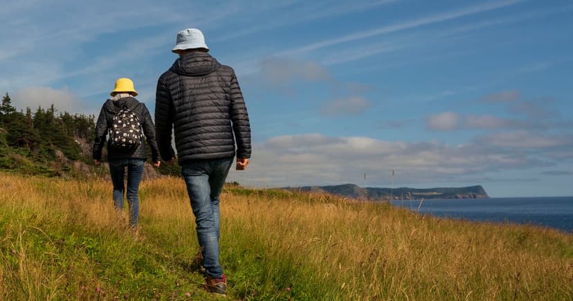 A couple walk one behind the other along a grassy coastal track above the sea, with a distant headland ahead of them.