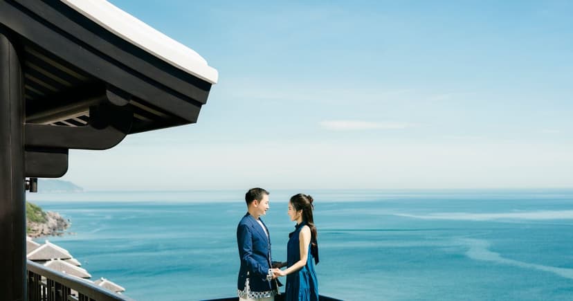 An elegant couple dressed in navy stand facing each other on a terrace high above a bright blue sea.