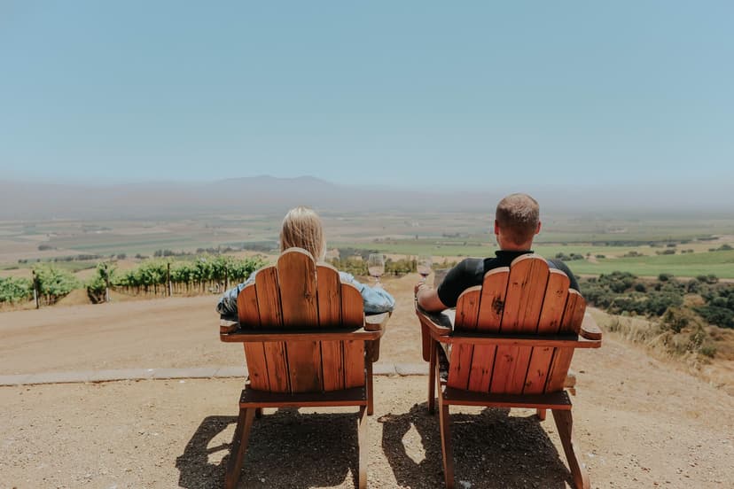 A couple sits side by side in wooden lookout chairs with glasses of wine, gazing out over green vineyard rows and a hazy summer valley