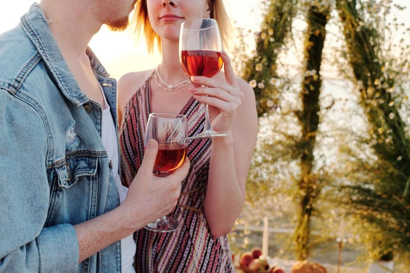A young couple in summer clothes stands close together at golden hour, holding two glasses of rosé in front of a flower-covered arch