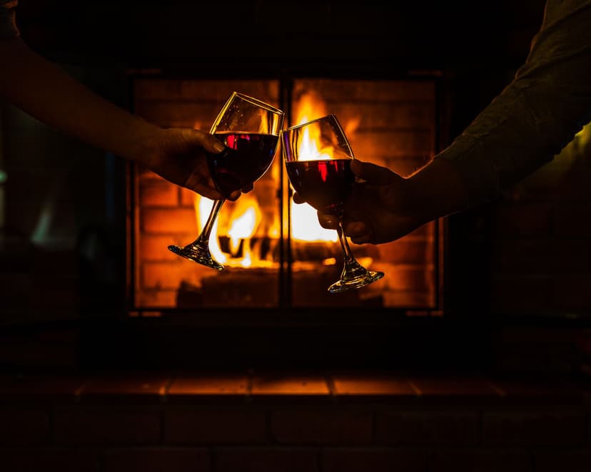 Two people toasting with glasses of red wine in front of a glowing fireplace, their hands and glasses silhouetted against the flames