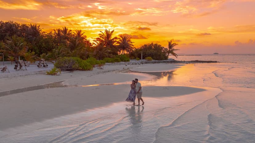 Couple strolling arm in arm across the wet sand of a tropical island beach at golden sunset, the woman in a flowing summer dress and the man in a light shirt, with palm trees silhouetted against the glowing sky behind them