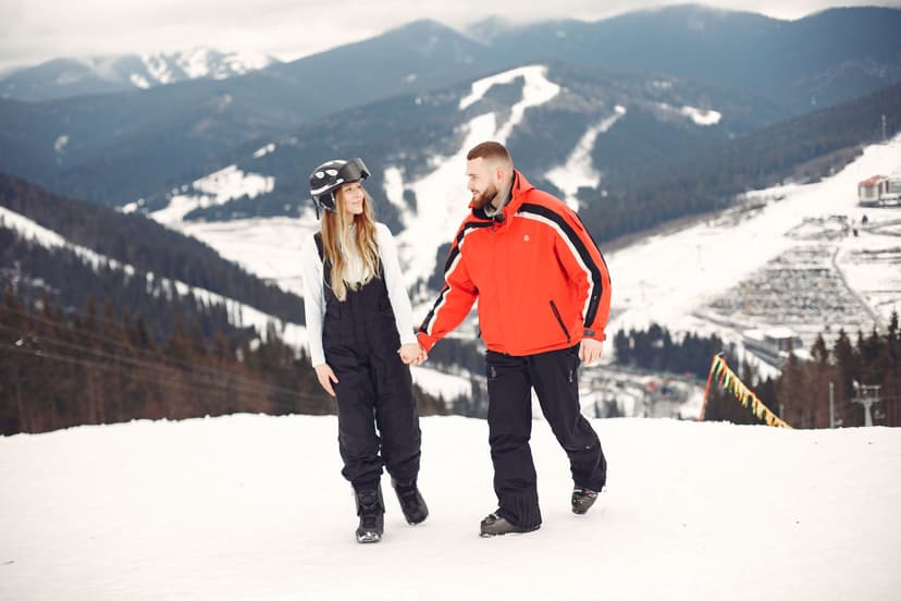 A smiling young couple in ski gear holding hands on a snowy slope, with ski runs, forest and a resort village spread across the mountains behind them