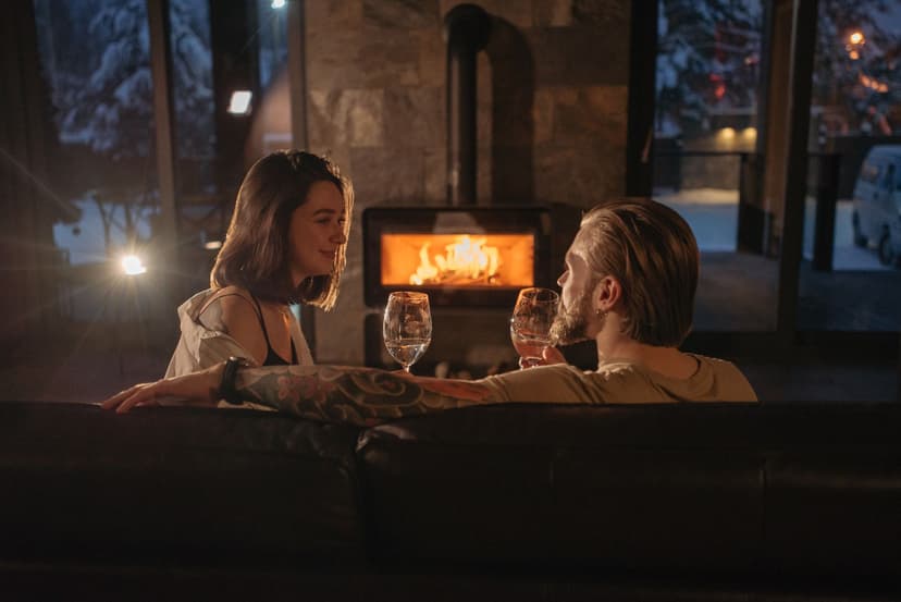 A couple sharing glasses of wine on a sofa beside a glowing fireplace, with snow-covered trees visible through the lodge windows at night