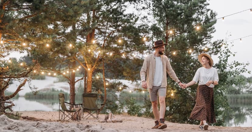 A man and woman in sun hats walk hand in hand past camp chairs and string lights beside a pine-lined lake.