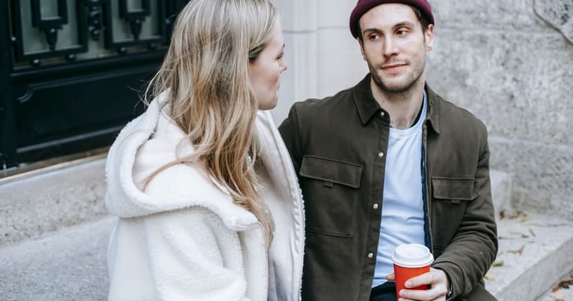 A woman in a white fleece and a man in a maroon beanie sit talking on stone steps, holding takeout coffee.