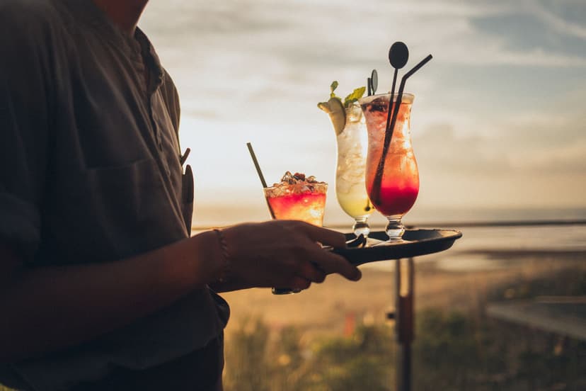 Ober met een dienblad met drie kleurrijke cocktails op een strandclub bij zonsondergang, met de zee wazig op de achtergrond