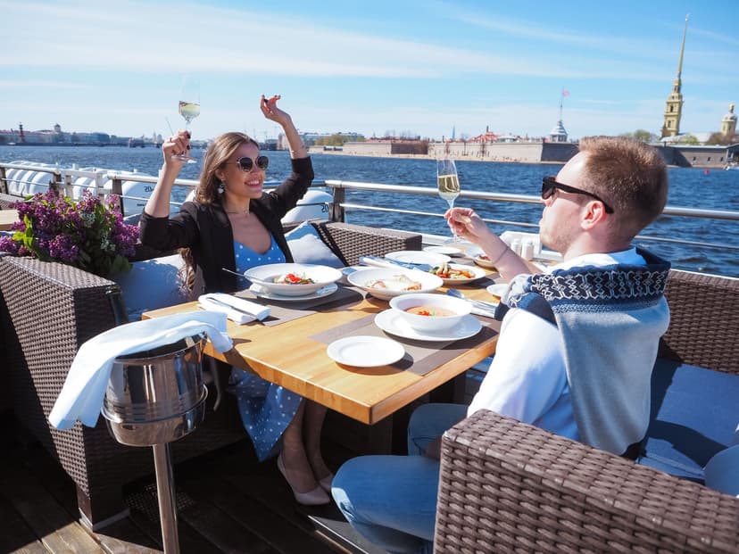 Velklædt par skåler i champagne over et veldækket bord på en restaurantterrasse ved vandet - kvinden i lyseblå sommerkjole og mørk blazer, manden i lys skjorte med sweater over skuldrene, og havneudsigt i baggrunden