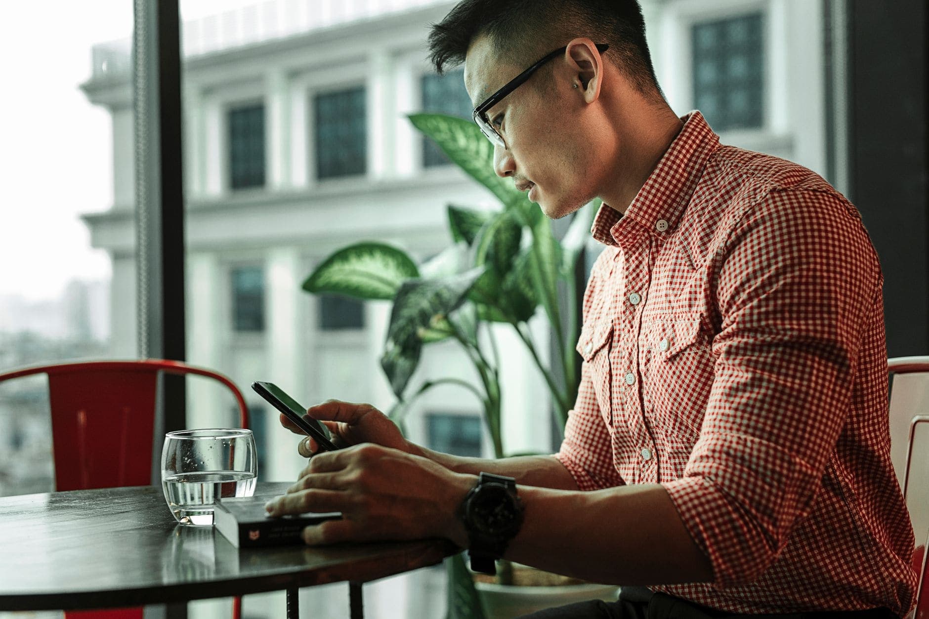A man in glasses and a red checked shirt reads his phone at a sunlit cafe table by a window
