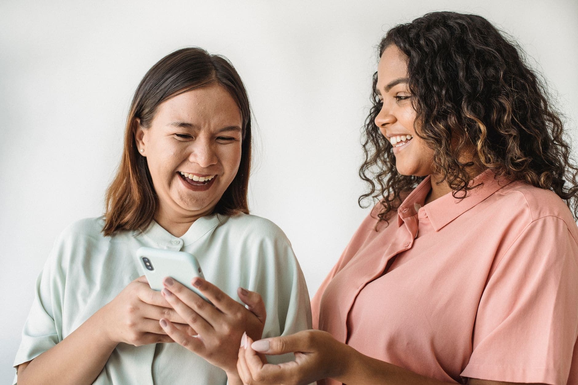Two women smile at a smartphone in front of a pale wall.