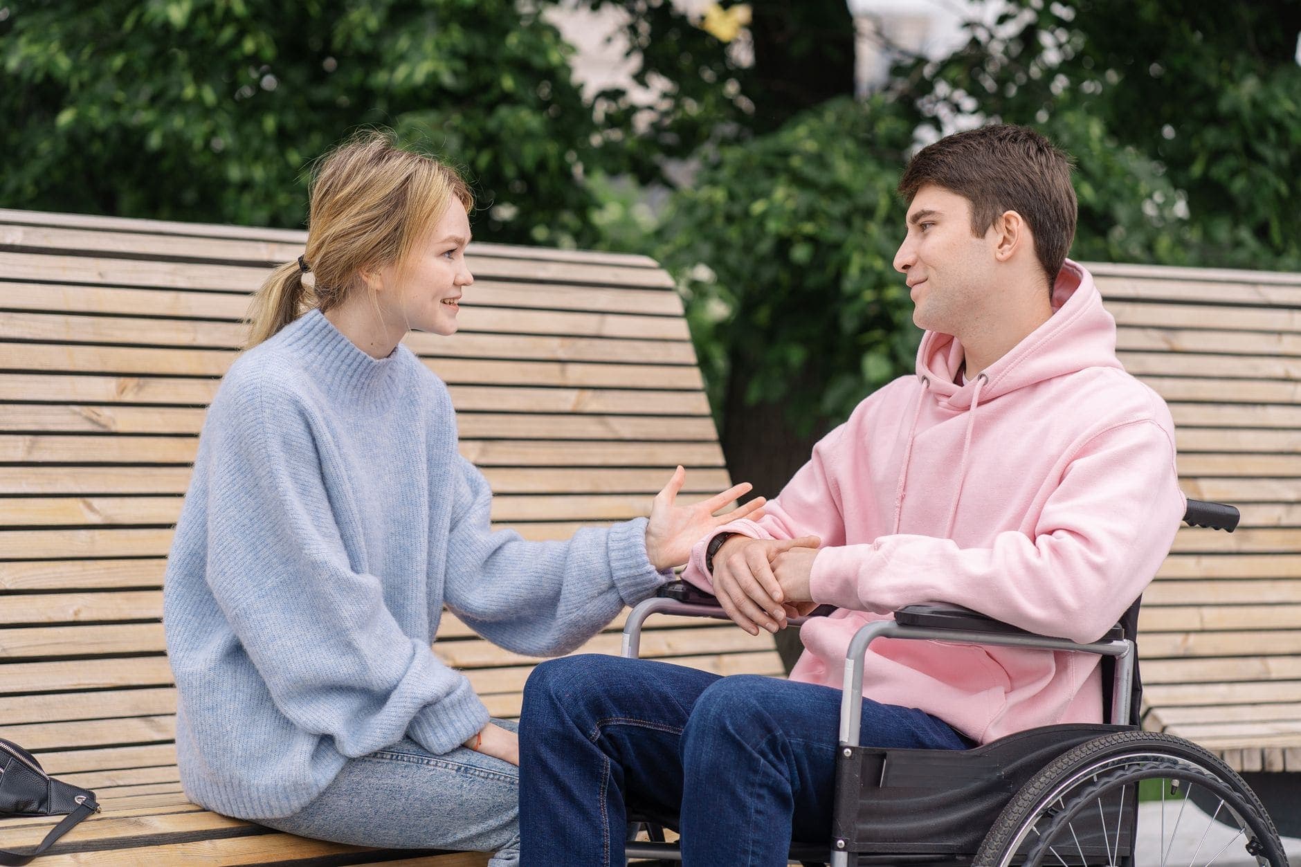 A woman in a blue jumper gestures towards a man in a pink hoodie beside a park bench.