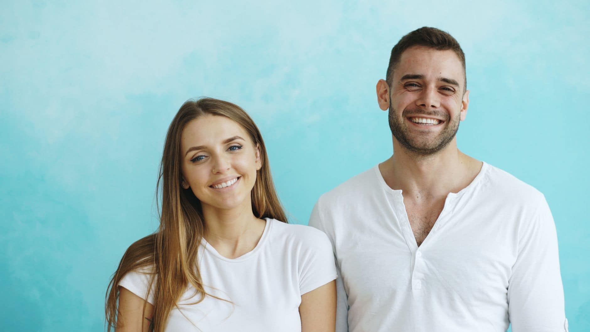Smiling young couple in white attire standing against a bright blue background.