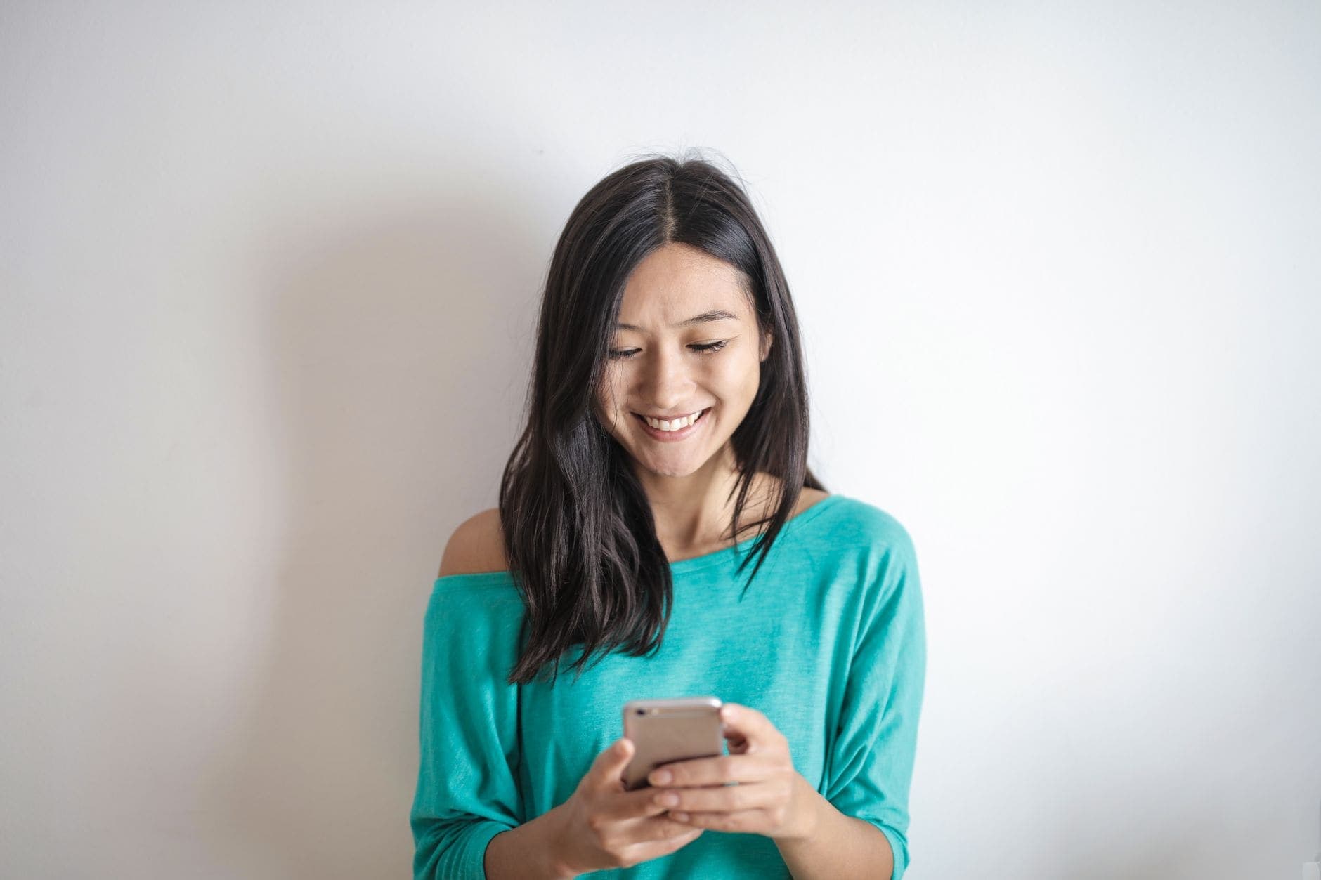 A woman in a turquoise top smiles while looking down at the phone in her hands.