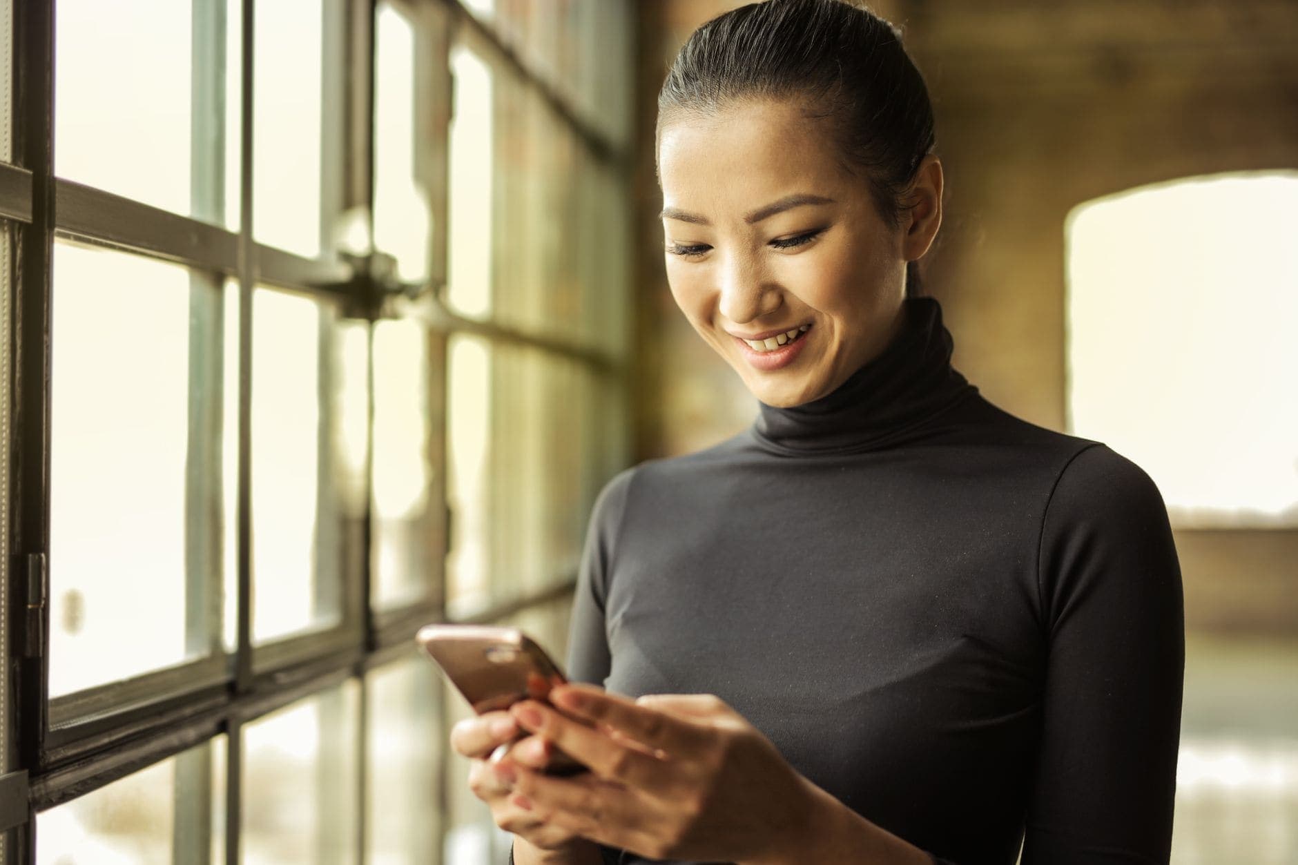 A woman in a black top smiles down at a phone beside a sunlit window.