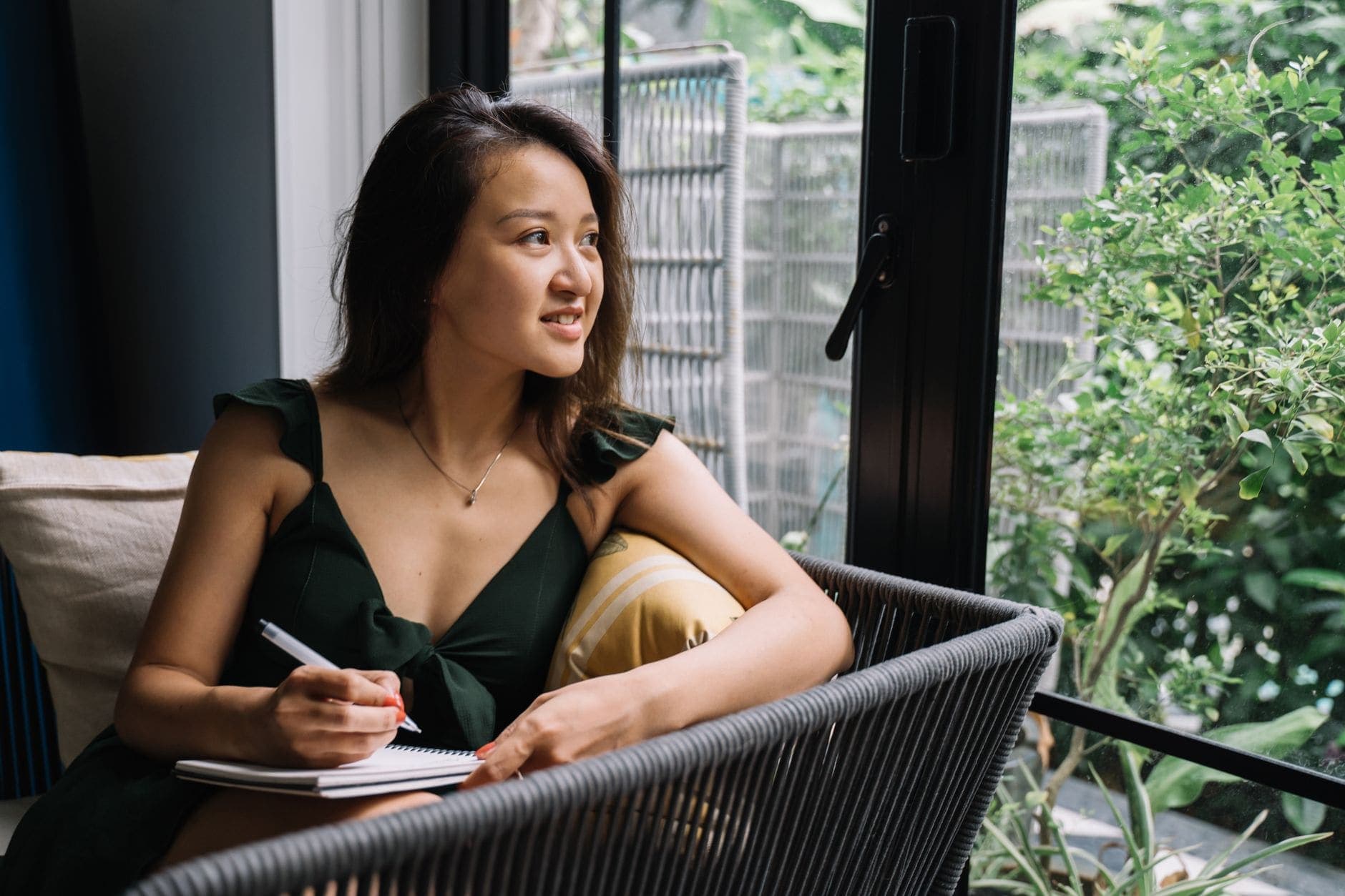 A woman in a green dress looks up from a notebook while holding a pen beside a window.