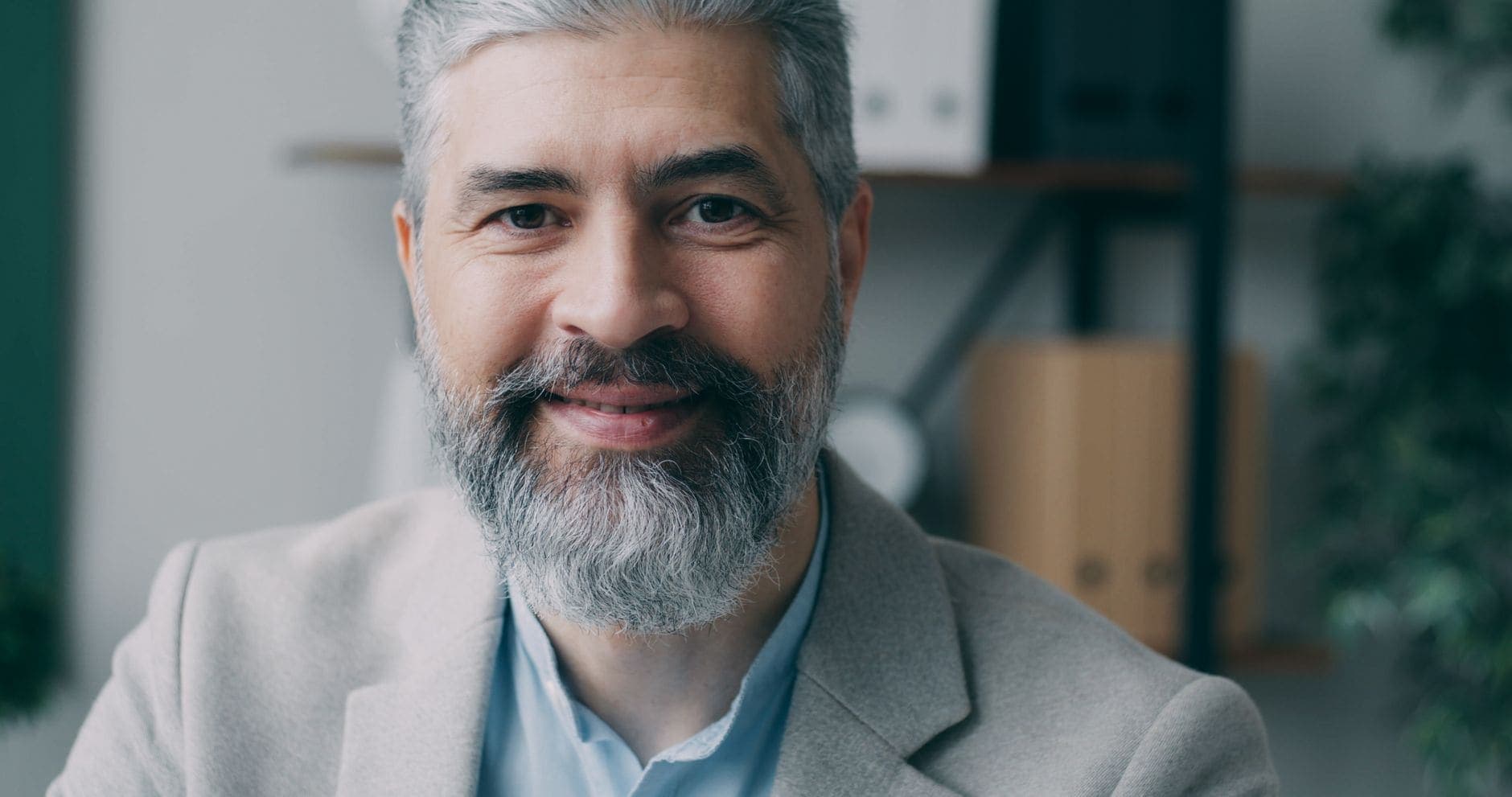 A grey-haired man with a beard smiles in a light jacket over a blue shirt.