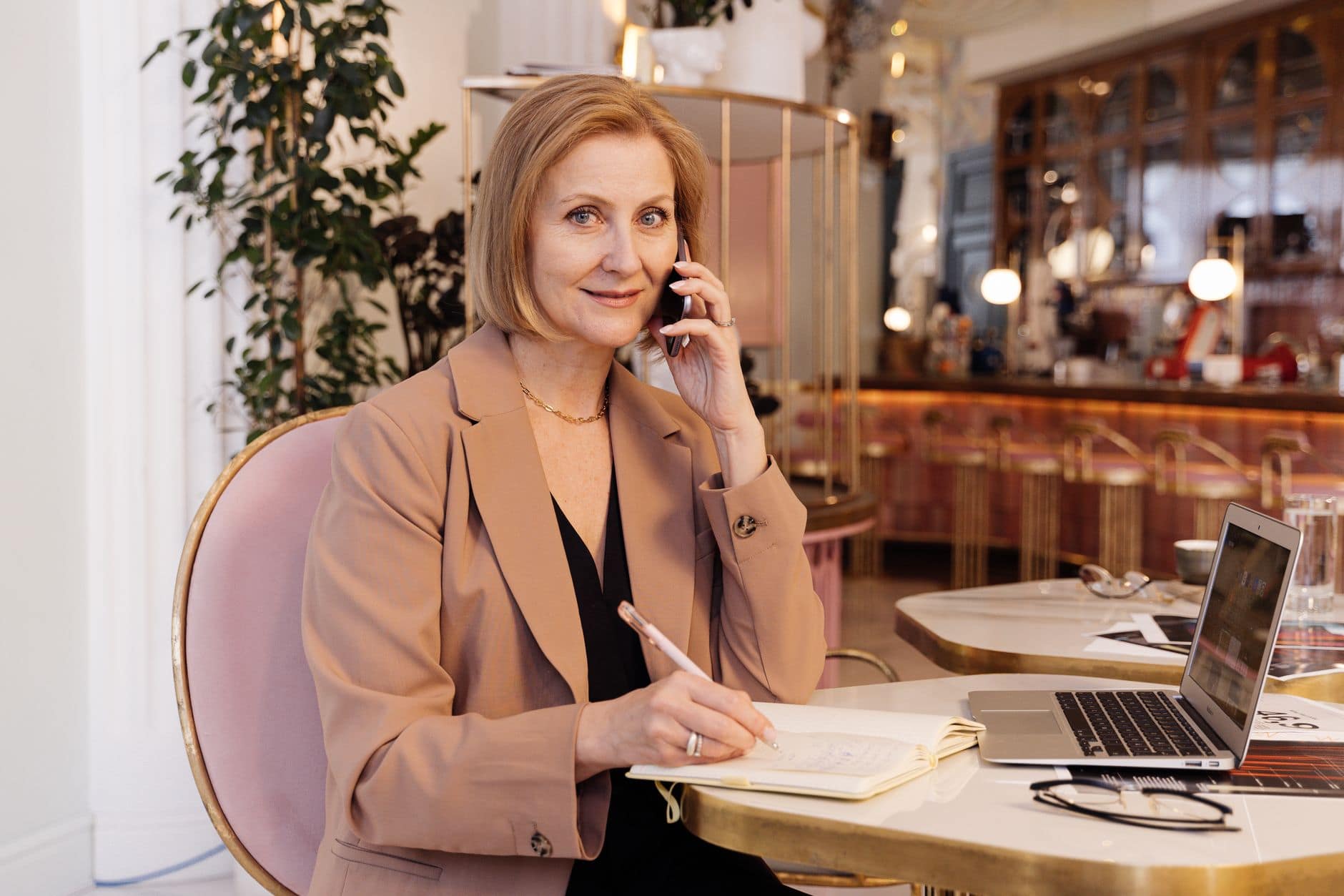 Une femme en blazer beige téléphone et écrit dans un carnet à une table de café.