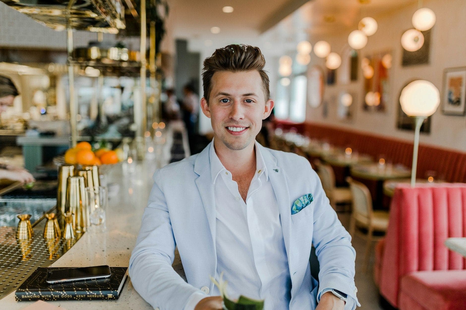 Un uomo sorridente in blazer azzurro e camicia bianca seduto al bancone di un ristorante luminoso.