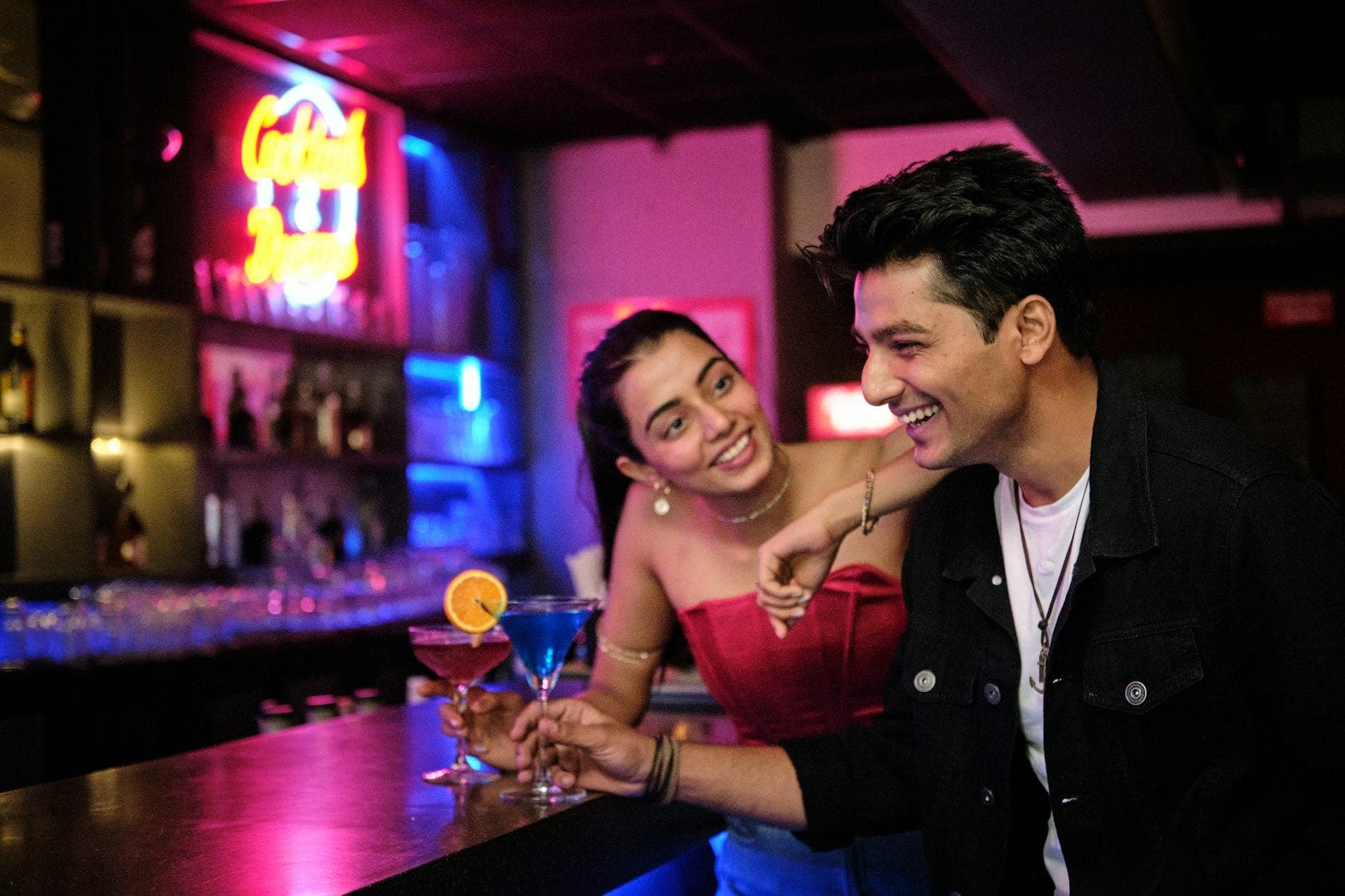 Pareja riendo en la barra de un bar con luces de neón, ella con un top rojo y un cóctel en la mano