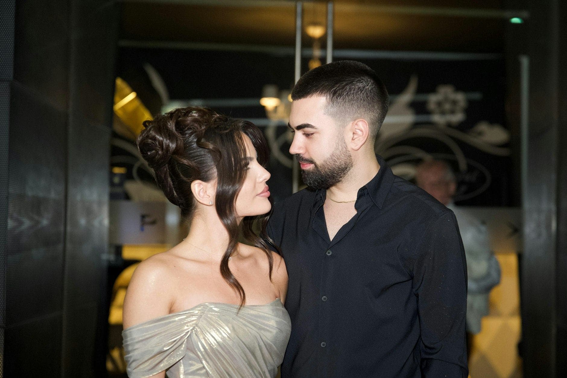 Žena s elegantným účesom v saténových šatách a bradatý muž v čiernej košeli sa na seba nežne pozerajú