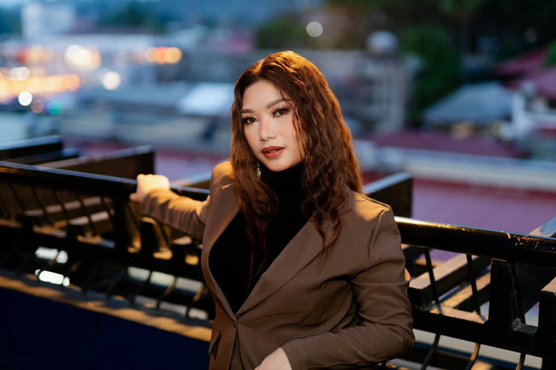 Une femme aux longs cheveux auburn, en blazer marron, sur un balcon dominant la ville au crépuscule.