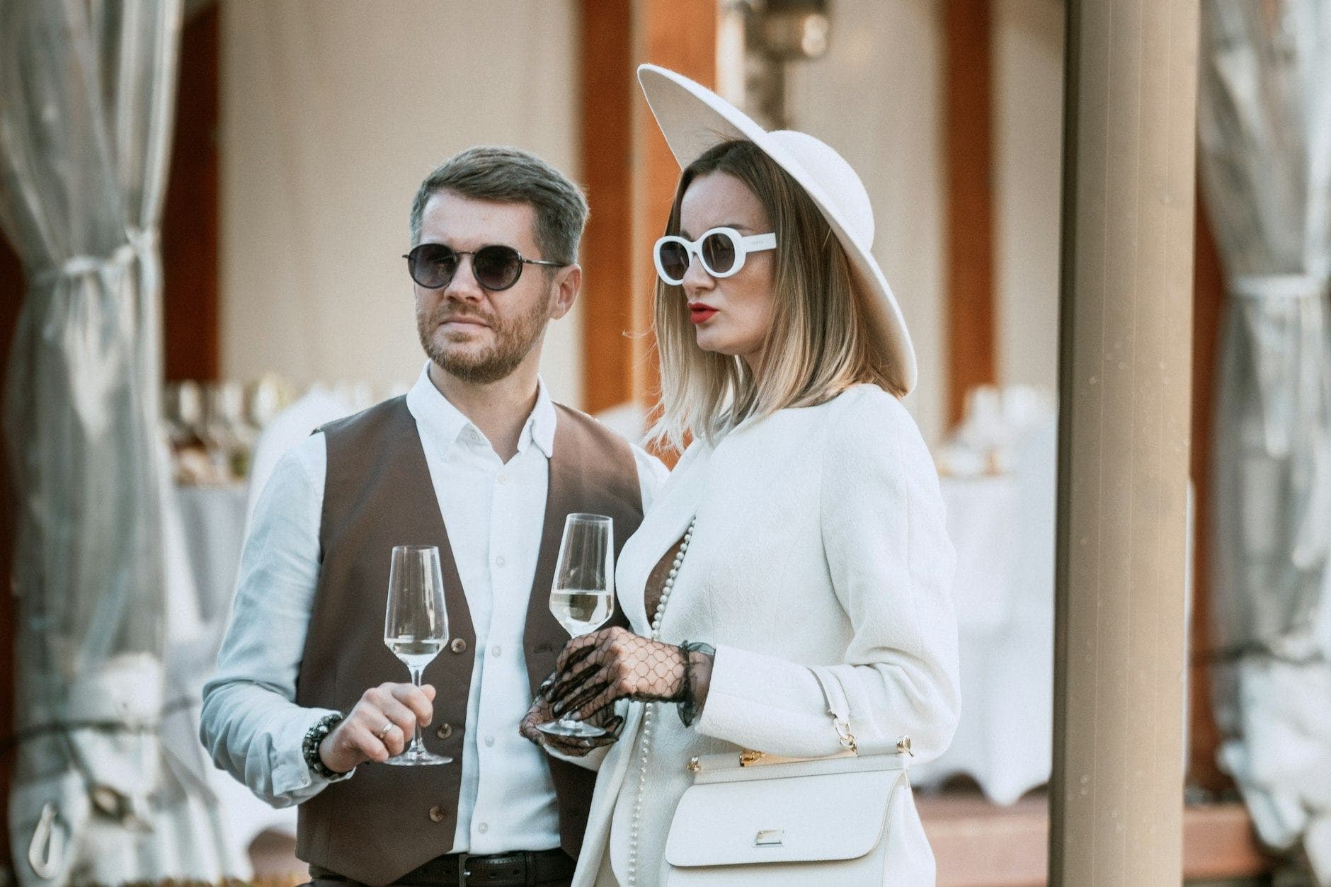 Un couple chic en lunettes de soleil, elle en blanc et chapeau, lui en gilet, verres de vin blanc à la main.