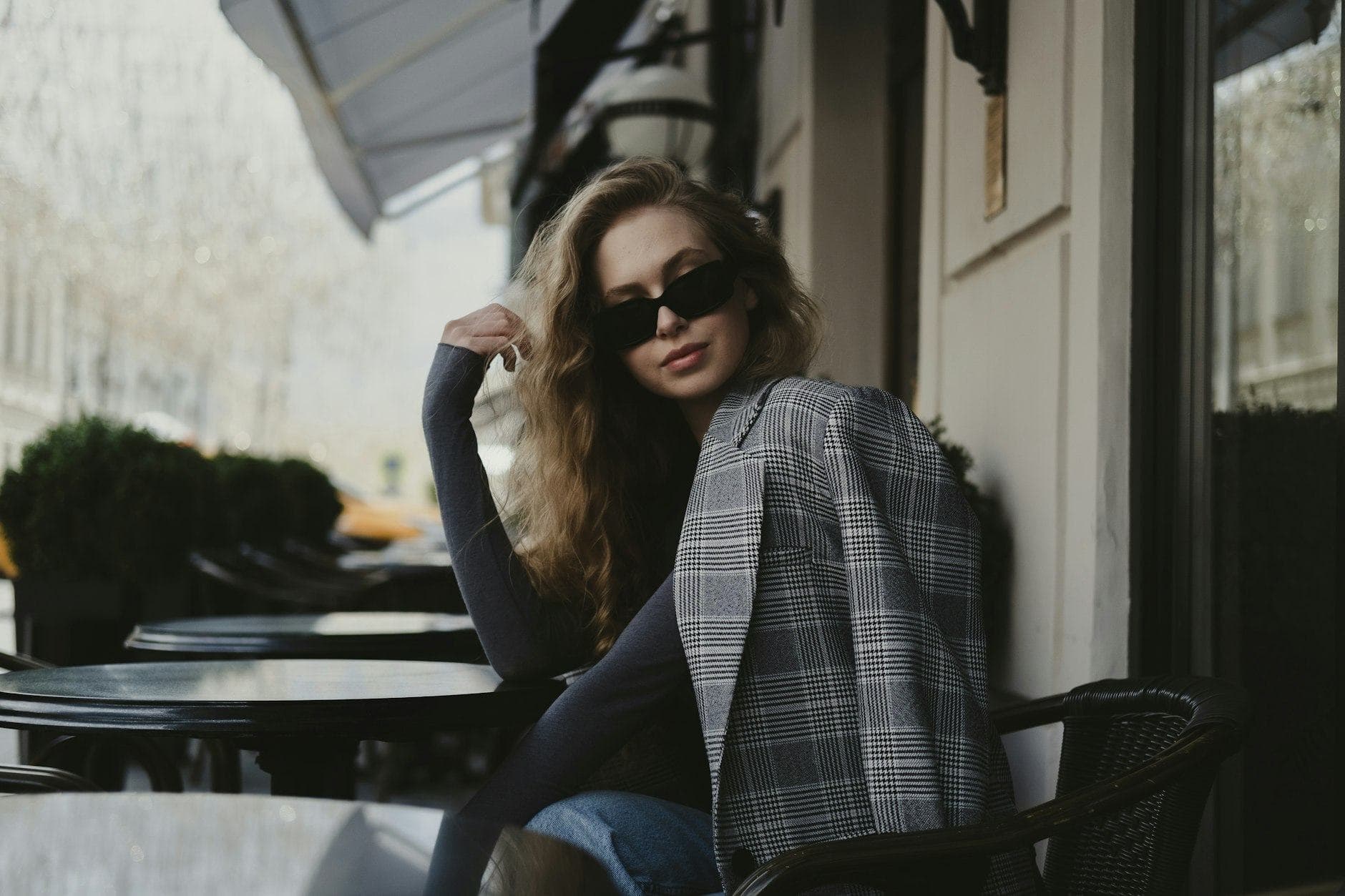 Une femme en lunettes de soleil et blazer à carreaux, assise à la terrasse d'un café devant un bâtiment
