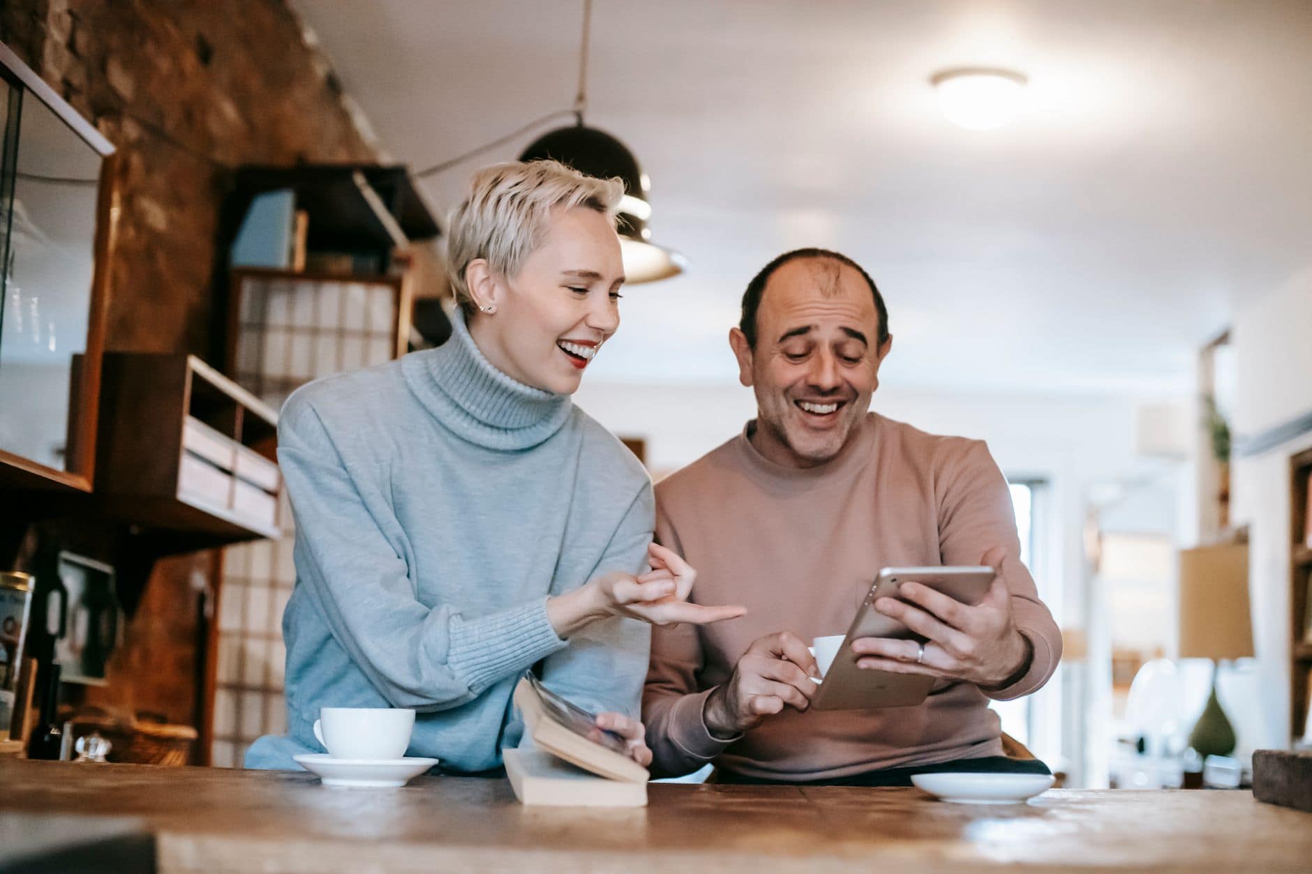Eine Frau mit kurzem blondem Haar und ein älterer Mann lachen gemeinsam über ein Tablet an einem Cafétisch.